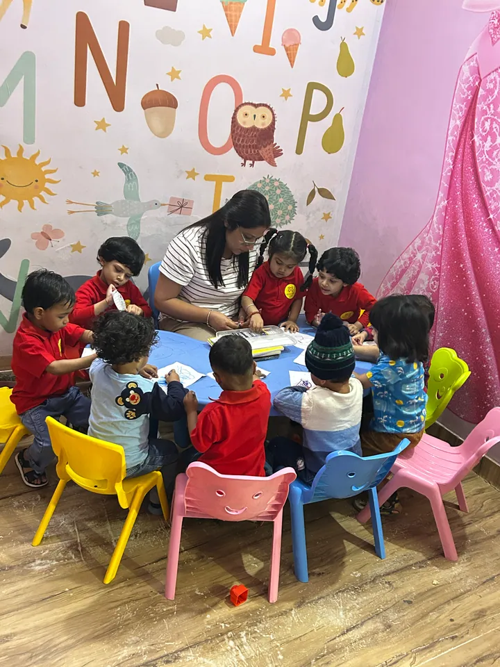 Poonam ma'am at the round table with a group of children under the alphabet mural