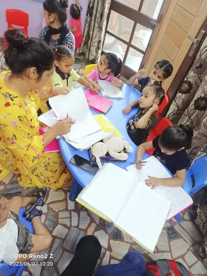 A teacher in yellow sits at the blue round table checking the children’s open notebooks