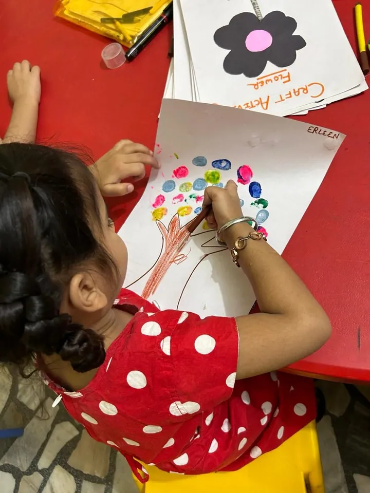 A girl in a red polka-dot dress presses coloured thumbprints onto a tree drawing