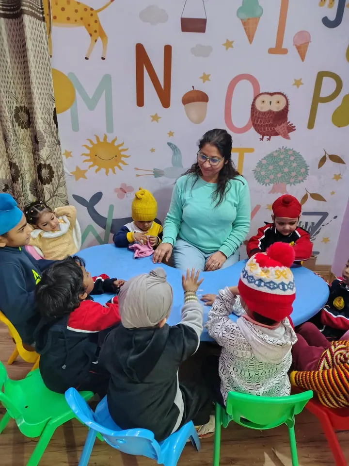 Poonam ma'am at the round table with a small group of children in woolly hats under the alphabet mural