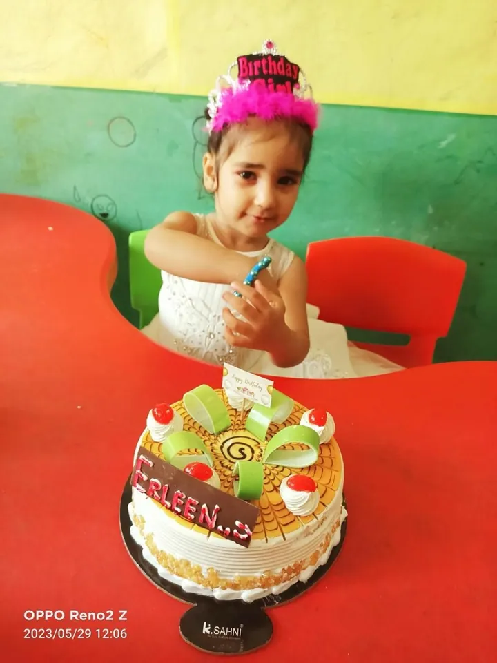 A girl in a birthday crown and white dress holds a candle beside her cake at the red table