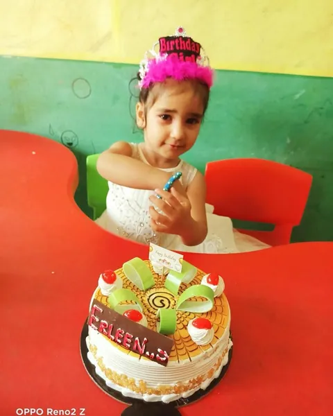 A girl in a birthday crown and white dress holds a candle beside her cake at the red table