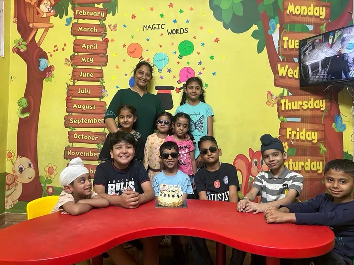 Children crowd round the red table with a birthday cake, in front of the months and magic words wall