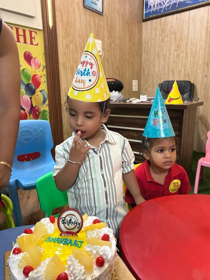 A boy in a party hat licks icing off his finger beside his cake, a friend in a party hat next to him