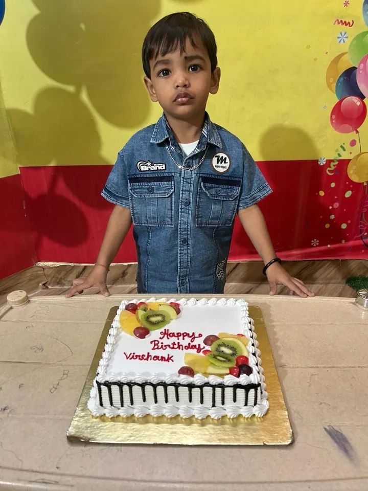 A little boy in a denim shirt stands behind his fruit-topped birthday cake