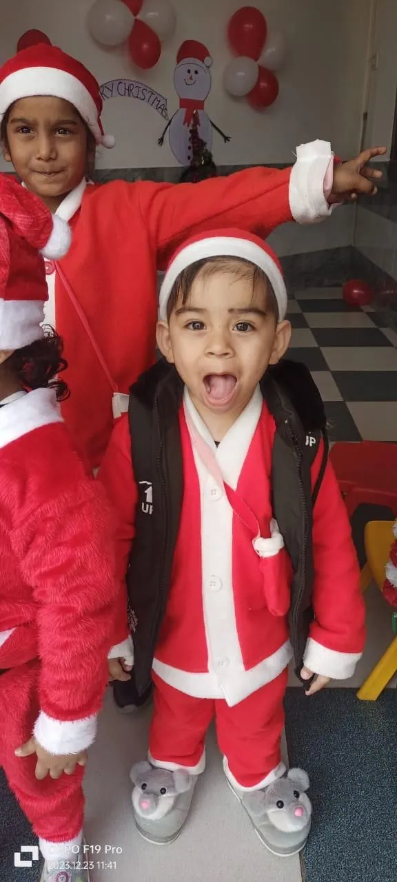 Two children in Santa suits, one shouting happily, by the Christmas decorations