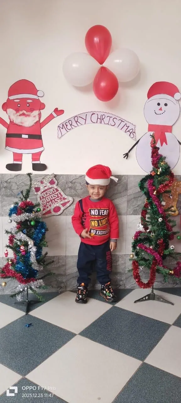 A little boy in a Santa hat stands in the Christmas corner between two decorated trees, under a Merry Christmas banner