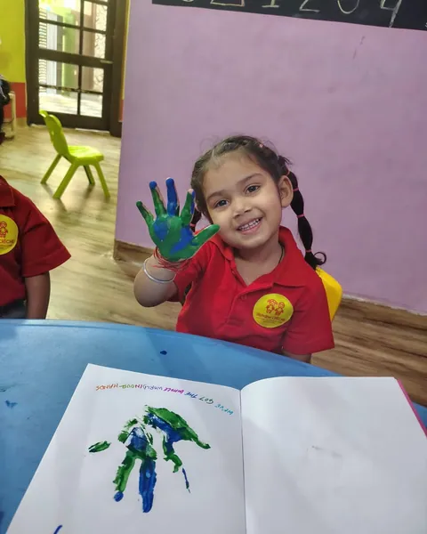 A girl in the red alphabet CRÈCHE uniform holds up her green and blue painted hand above her handprint page