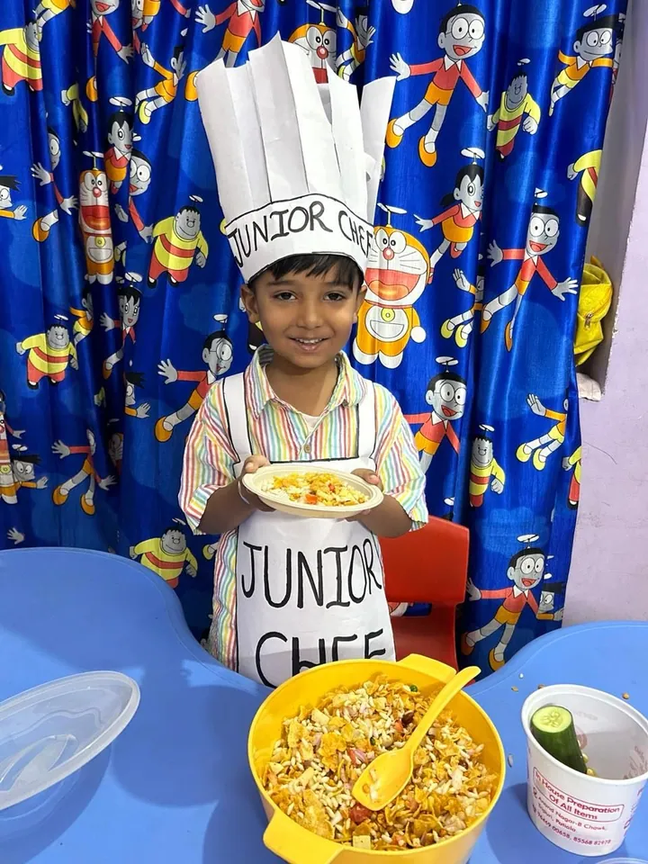 A boy in a paper Junior Chef hat and apron holds up his plate