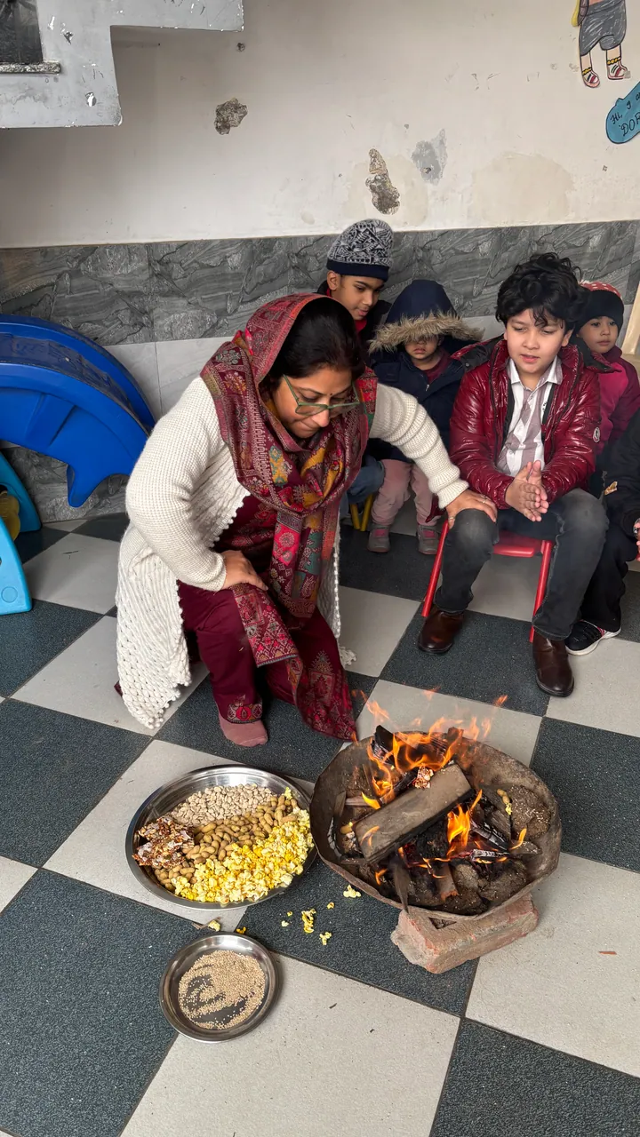 Poonam ma'am kneels by the Lohri fire in the playing area as children watch