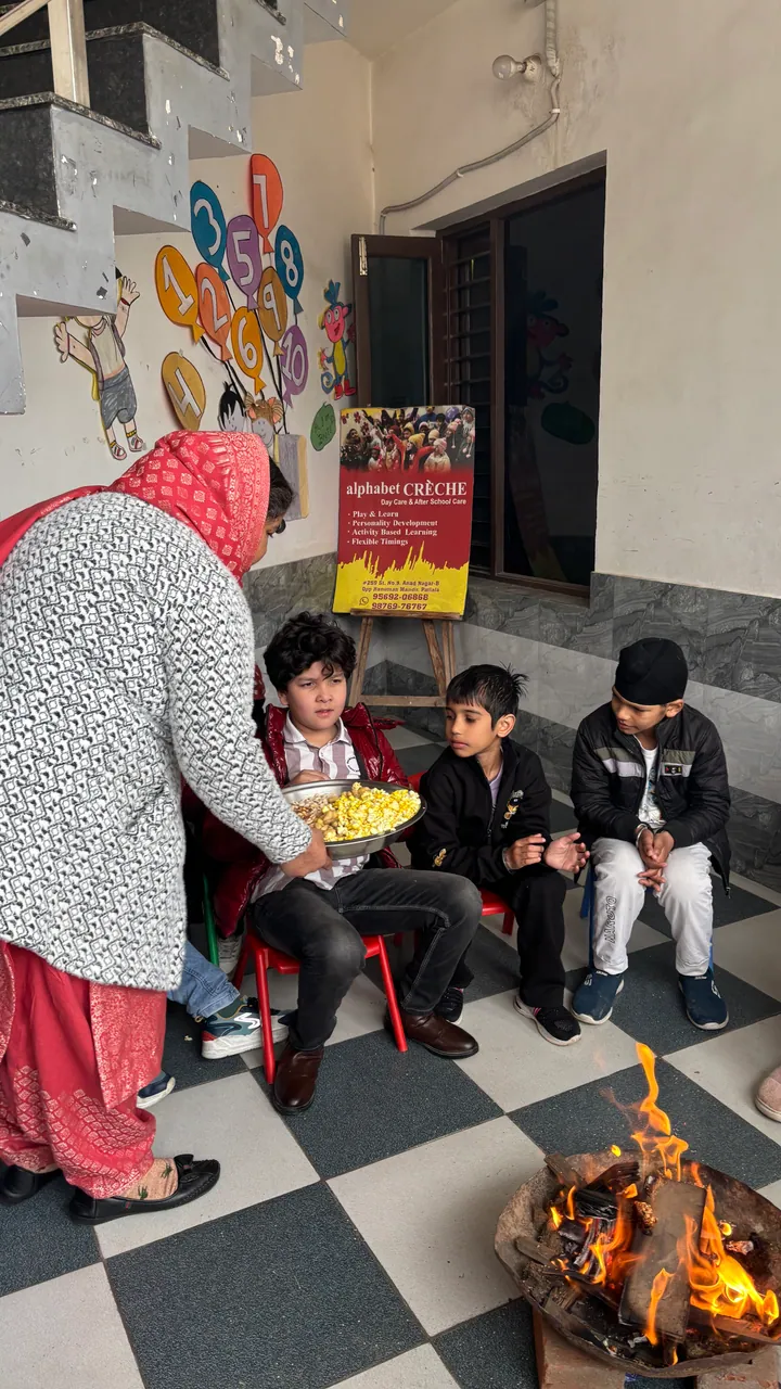 Manjeet ma'am holds a plate of popcorn out to seated children beside the Lohri fire