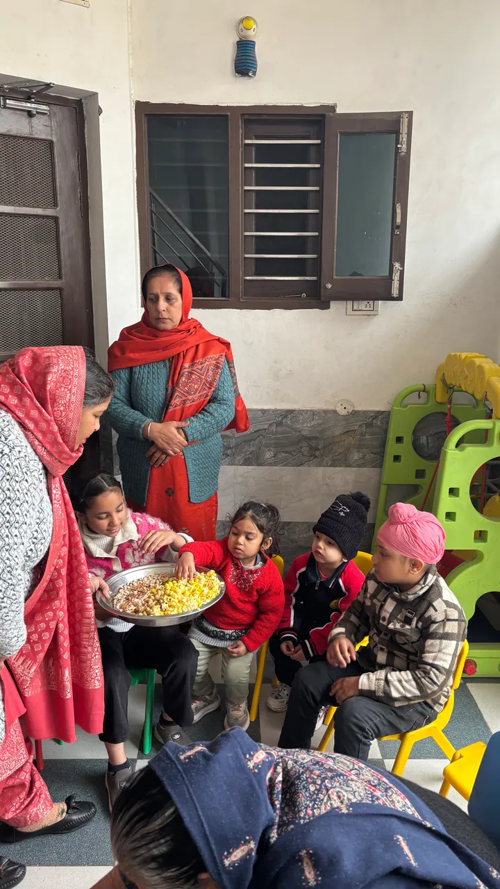 Indrajit ma'am stands at the back as the plate of popcorn goes round the children at Lohri