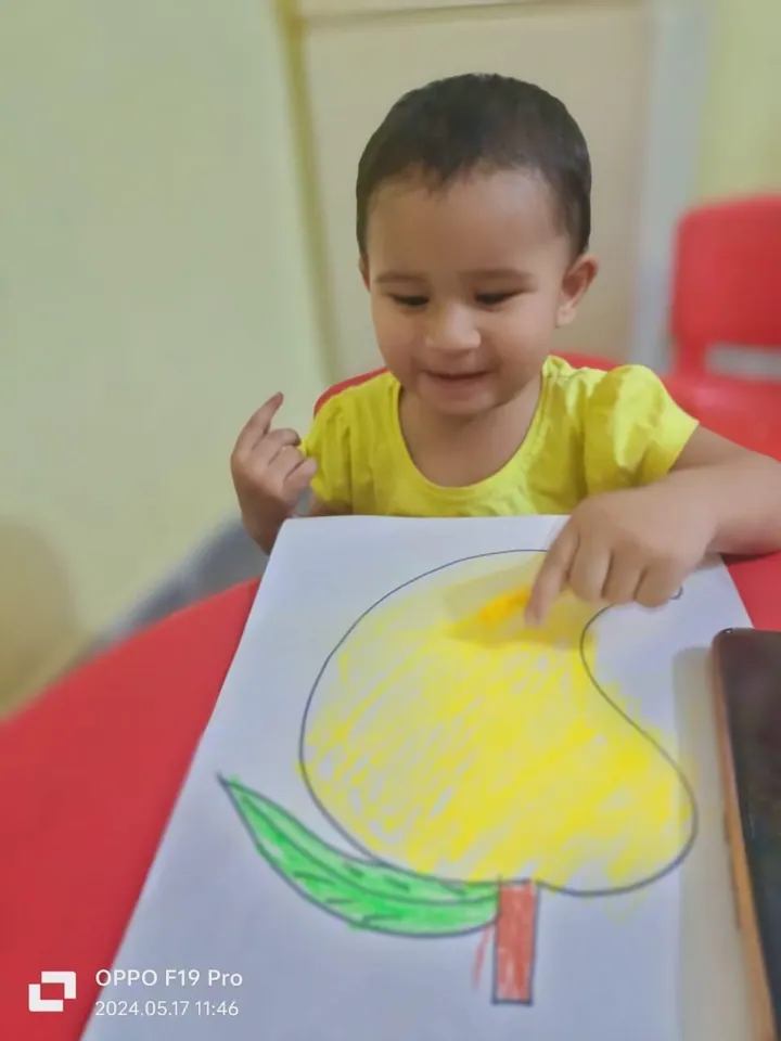 A smiling toddler colours a big mango outline yellow at the red table