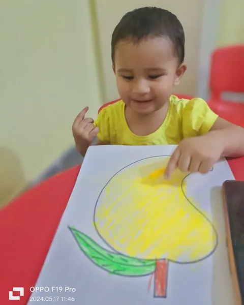 A smiling toddler colours a big mango outline yellow at the red table