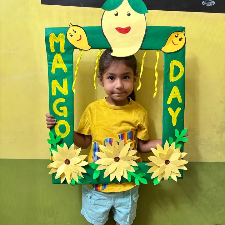 A girl in a yellow T-shirt holds up the green Mango Day photo frame with a smiling mango on top