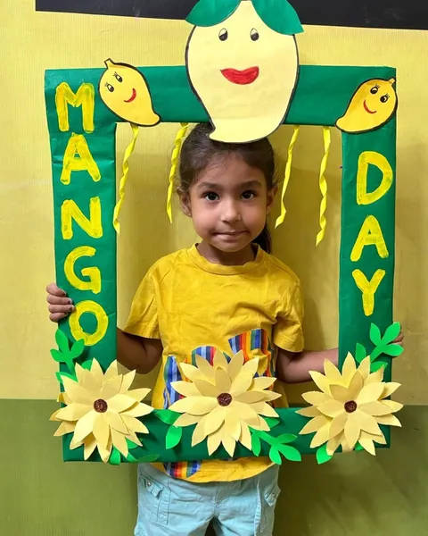 A girl in a yellow T-shirt holds up the green Mango Day photo frame with a smiling mango on top