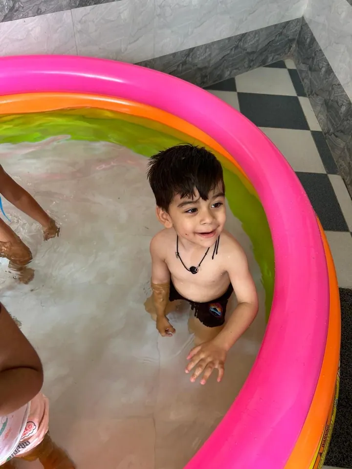 A little boy grins up from a pink paddling pool
