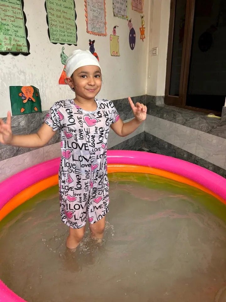 A girl in a patterned swimsuit stands in the paddling pool, pointing up and smiling